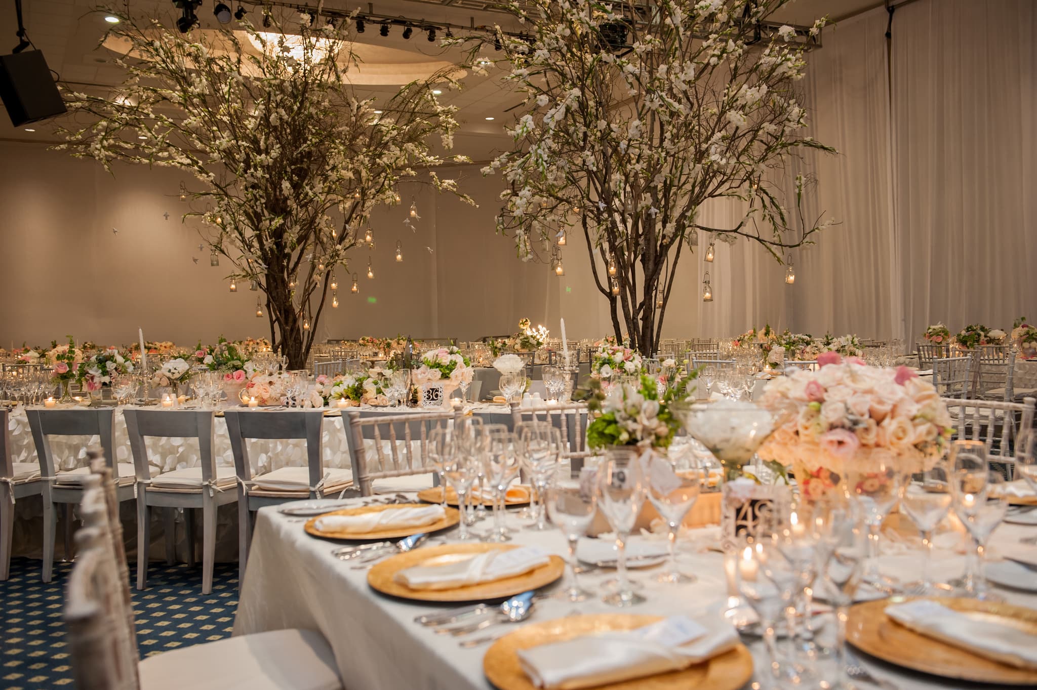 Banquete de boda con mesas largas, platos base dorados y árboles florales con velas colgantes